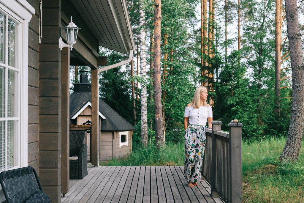 Our cottage via Mokkiavain Cottages. A girl stood on the veranda of a log cabin, in a woodland in Tampere.